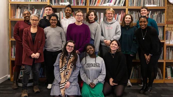 A group of people stand in a row in front of a bookshelf with three people kneeling on the floor in front, they are all facing forward to the camera smiling