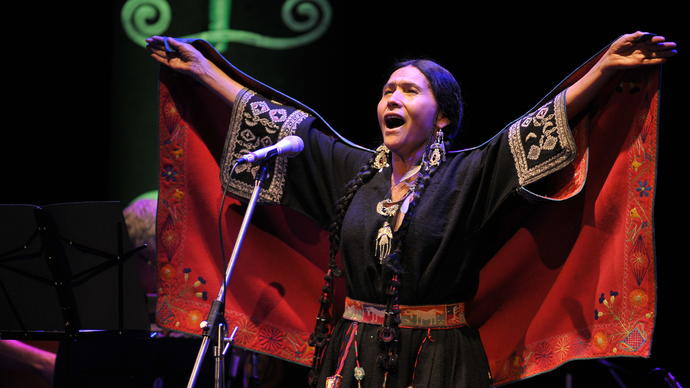 Luzmila Carpio singing, she is wearing a colourful traditional indigenous outfit with her hands outstretched above her