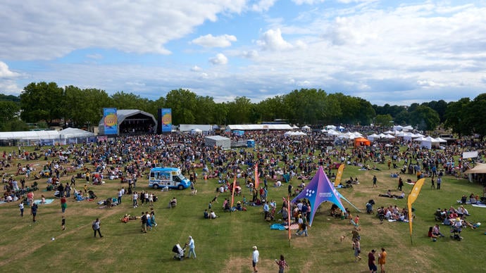 A high up photo of a field with lots of people standing and sitting on it with a large music stage in the background