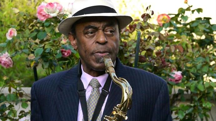 colour photo of archie shepp holding a saxophone standing in front of a rose bush
