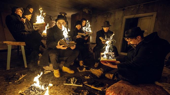 A group of people dressed in black sit in a decaying building holding books that burn in their hands.