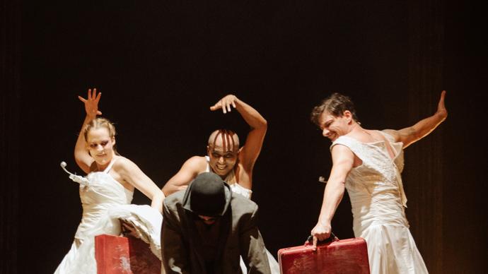 Three performers in white dresses appear to be dancing or moving behind a figure in a coat and hat in front of them. They each carry a red suitcase. 