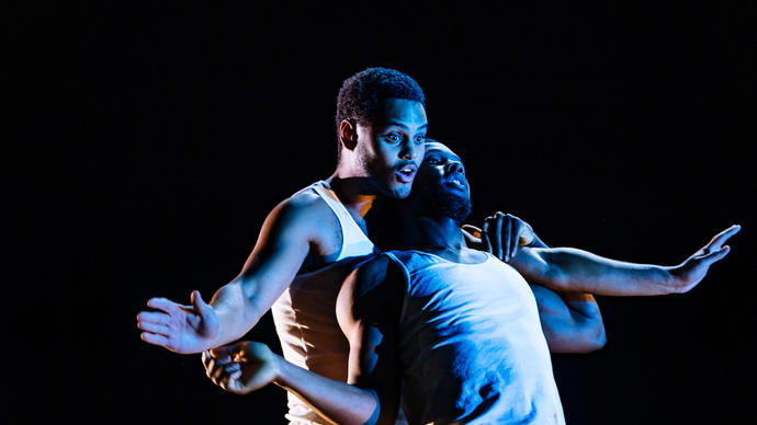 A young man holds another in his arms, they are both dressed in white. Behind them, other people in the company watch on, intrigued.