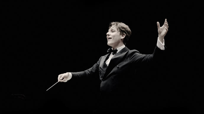 Klaus Makela is conducting in front of a black background, his right hand holding the baton and his left hand above his shoulder