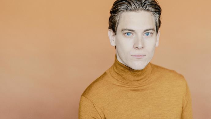 Klaus Makela looks at the camera, in an orange jumper in front of an orange background