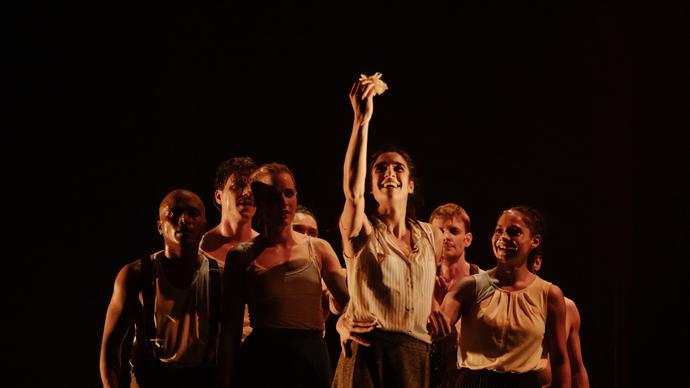 A production photo of the Gecko Theatre ensemble performing The Wedding. A performer stands holding a flower in the air. Behind them, a crowd of performers watch.