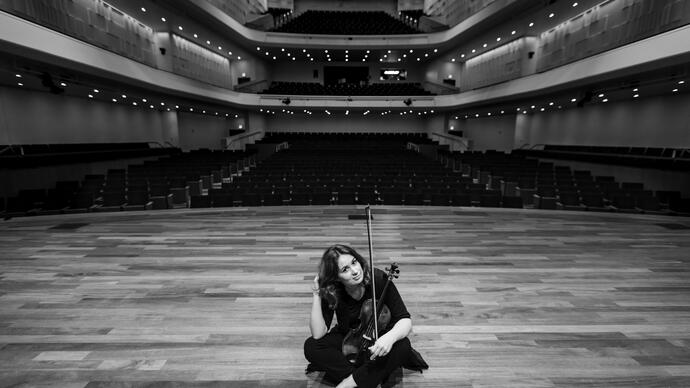 Patricia Kopatchinskaja sitting on a stage holding her violin in her lap