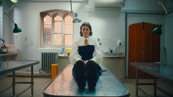 A person sits on a morgue table with their hands in their lap looking at the camera.