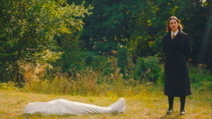 A person stands in a field, next to a corpse covered in tarpaulin.