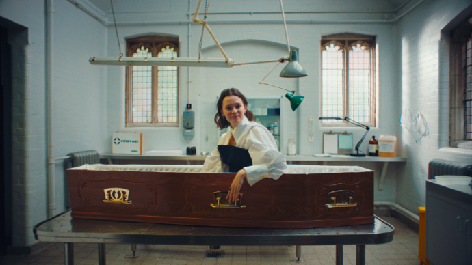 Person sits upright in a coffin, with the camera facing them side on.