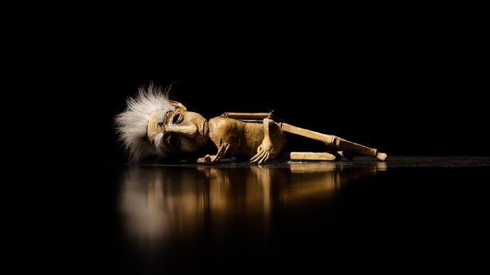 A puppet lies naked on the floor in darkness. 