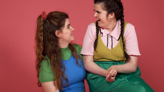 A lady with brown hair, wearing green and blue layers is smiling looking towards another lady who is laughing back at her. The lady on the right also has brown hair that is tied into two plaits. She is wearing a pink short-sleeved top with a yellow layer on top and green trousers.