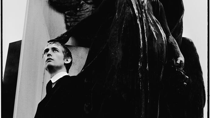 Black and white image of Neil Hannon posing beneath a statue 