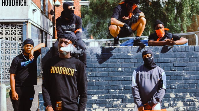 A group of performers in Hoodrich clothing, some wearing face coverings, sit and stand around a wall talking and smoking.