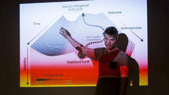 Playwright David Finnigan stands in front of a projector and speaks to the audience. On the screen, there is a slide about glacial-interglacial cycles.