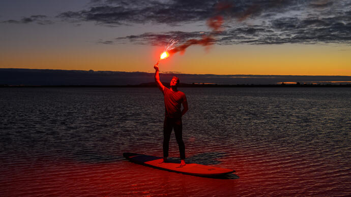 David Finnigan holds a flare and stands on a surf board in the ocean. It is sunset.
