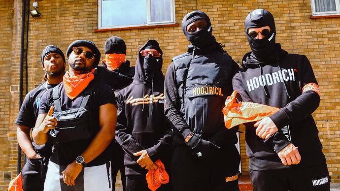 The ensemble dressed in black, some with face coverings, stand in front of some flats and look down the camera.