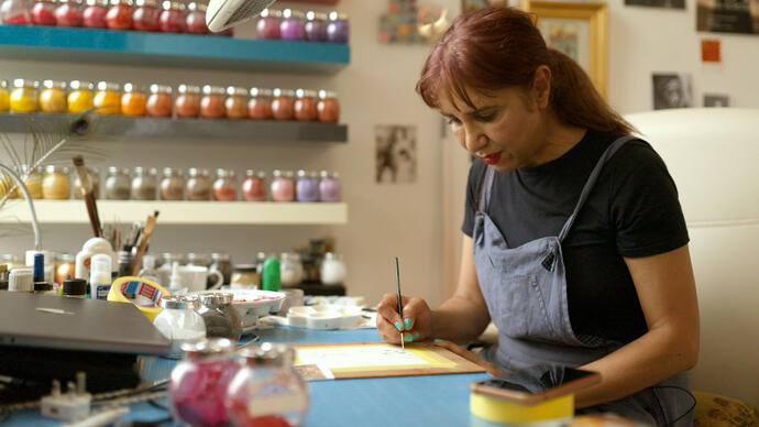 Artist Soheila is standing at her workstation in a blue apron and a paintbrush in her hand. On the wall behind are shelves with paint bottles.