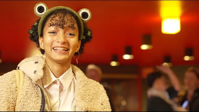 An audience member wears a headband with googly eyes on it and smiles at the camera whilst doing an interview about the show having just watched it.