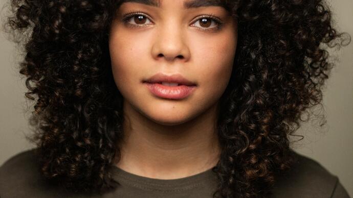 A headshot of Lydia West. They have black curly hair and brown eyes. They wear a black jumper and stare directly at the camera. 