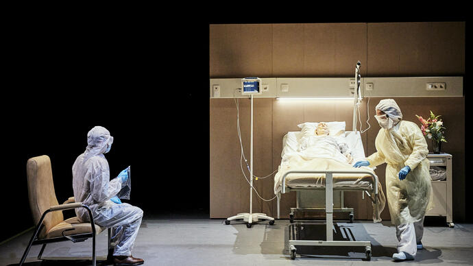 A person in a prosthetic face mask is lying in a hospital bed with two hospital workers around him wearing full PPC.