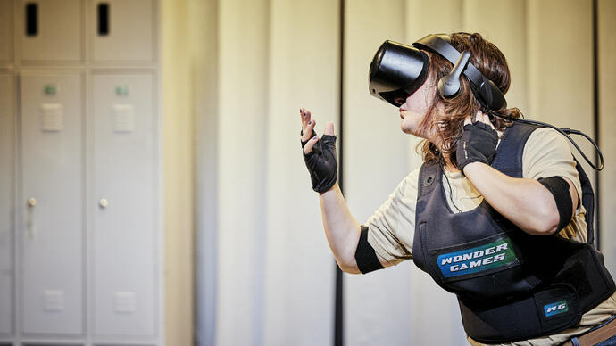 A white lady with long brown hair is wearing a VR headset and a body vest that reads 'Wonder Games'. They have one hand on their neck and another outstretched in front of them.