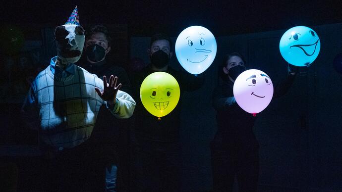 Bill the puppet stands in a line with four balloons. They are in darkness. The balloons have faces drawn on them.