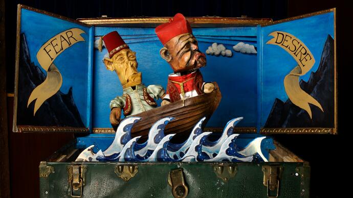 Two puppets in a travel case appear to be sailing on the sea in a boat. The words fear and desire are painted onto the back of the travel case on either side of them.
