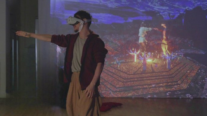 A dancer and choreographer stands in front of a large projection screen with a VR headset on. On the screen, they appear again in a game with another performer inside a digital 3d world.