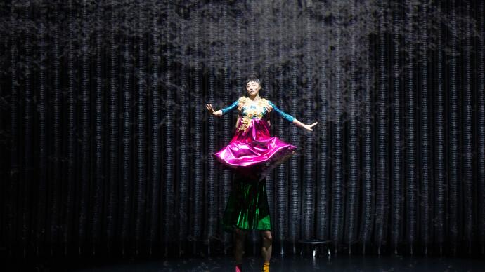 One performer stands on top of the others' shoulders to create one long person wearing brightly coloured clothing.