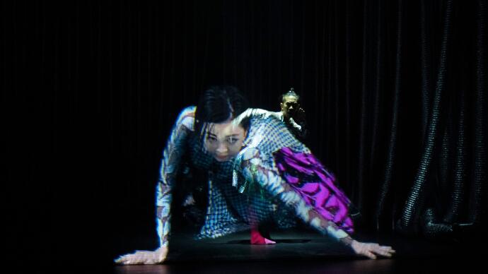 Two performers seem to be merged together while dancing on a stage. One looks like a hologram.
