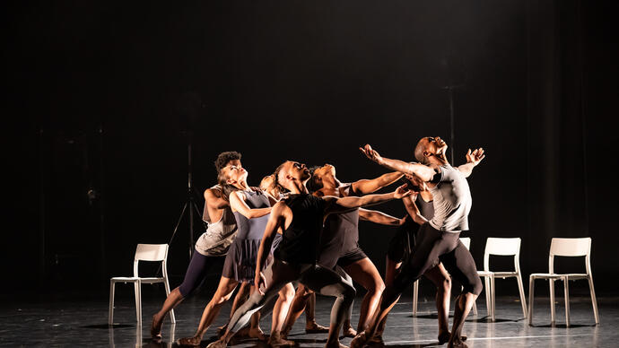 A group of dancers gather and seem to be pushing one dancer (whose arms are outstretched) ahead of them.