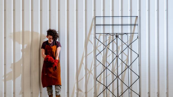 Artist Rachel Mars stands against a cream wall wearing protective clothing. A metal structure is next to them.