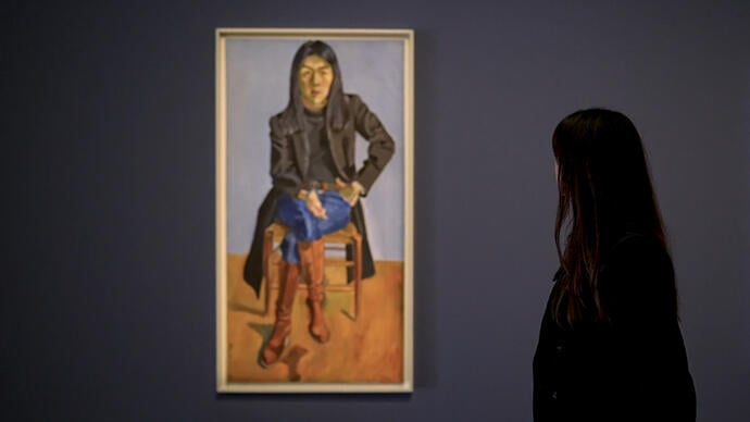 A person stands with their back to camera looking at a hanging portrait painting.