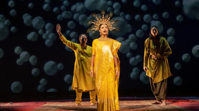 A performer wearing a gold crown sings, while two other performers stand behind.