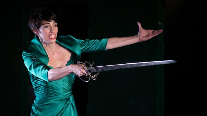 A performer wearing a green dress and holding a sword appears to be threatening someone. 