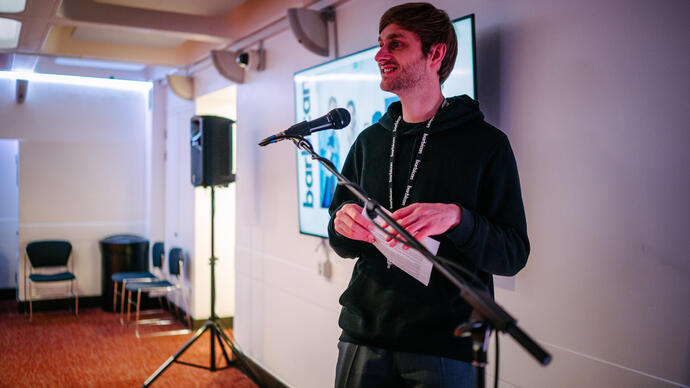 man wearing black hoodie with a lanyard around his neck, speaking into a microphone