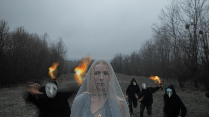 A person wearing a grey veil stands on grass holding a miniature carousel while a group of people dressed in black and wearing haunting facemasks run forwards holding fire.