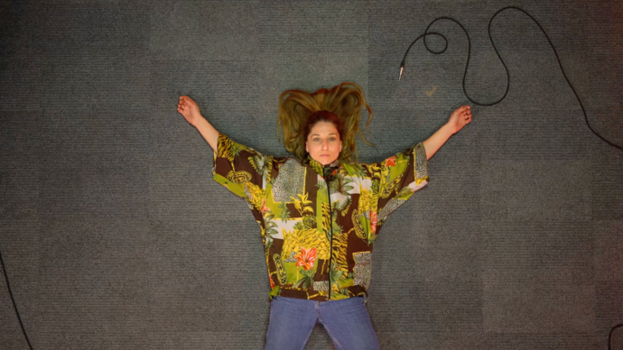 A dancer lies on the floor wearing a colourful top. Their arms are outstretched. 