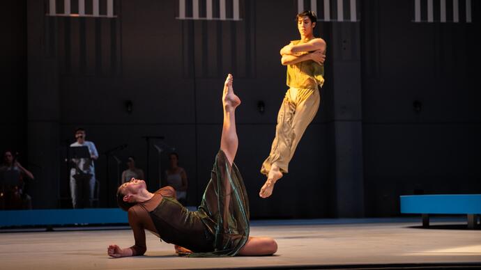 One dancer lies on the floor with their leg and toes pointing upwards while another jumps in the air behind them.