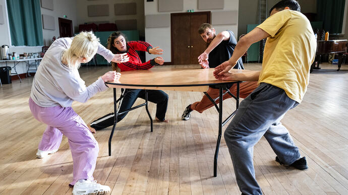Four people rehearse movement around a table, they bend to their sides dramatically.