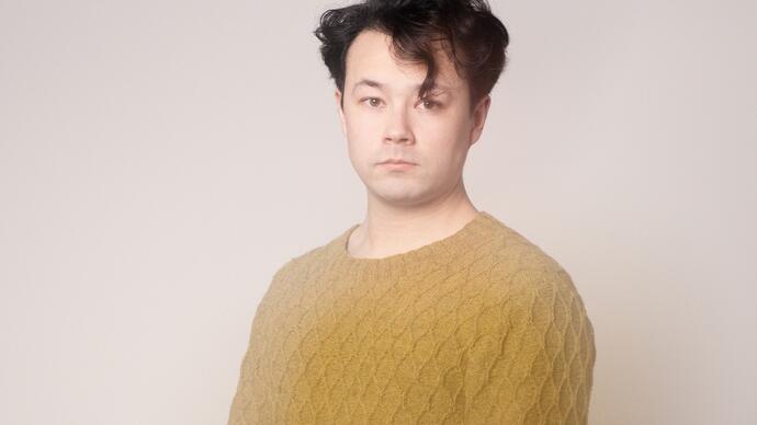 Photo of Sean Shibe wearing a mustard coloured jumper, standing in front of a white background and looking at the camera