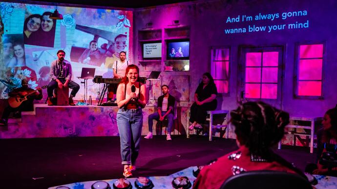 Rachel sits at her tech desk directing the show while the ensemble perform on stage, a person is lifted up in a chair.
