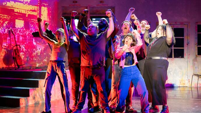 Members of the ZooCo ensemble perform on stage. They huddle in a group with their arms in the air, singing.