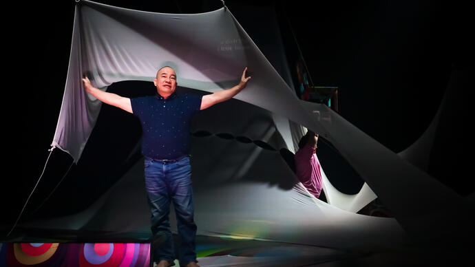 A person performs underneath a large piece of white draping fabric, they hold their arms out wide.