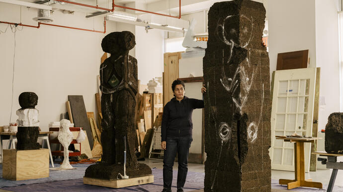 Huma Bhabha standing in a studio with two tall sculptures, one carved out and one with markings ready to be carved.