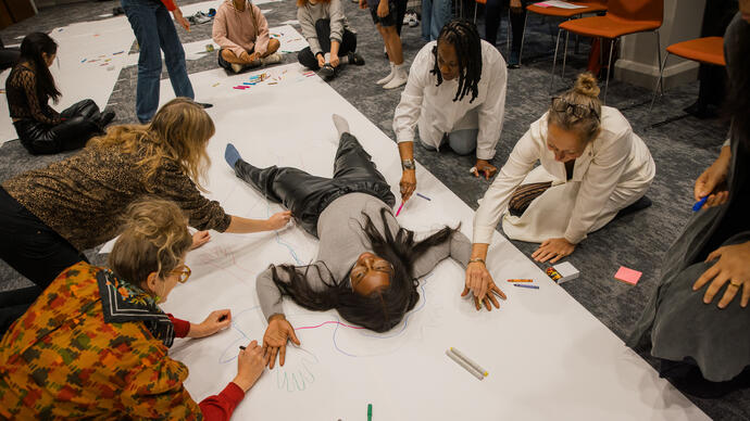 A person lies on the floor on a large piece of white paper. A group of people draw around them using a range of pens and pencils.