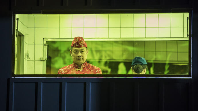 An Asian man in an enclosed kitchen-like space with a yellow-green background