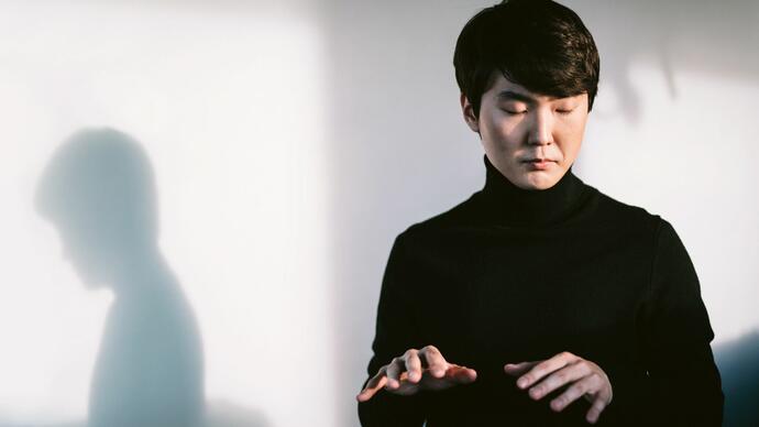 Pianist Seong-Jin Cho in front of a white wall, with eyes closed lifting his hands as if playing the piano