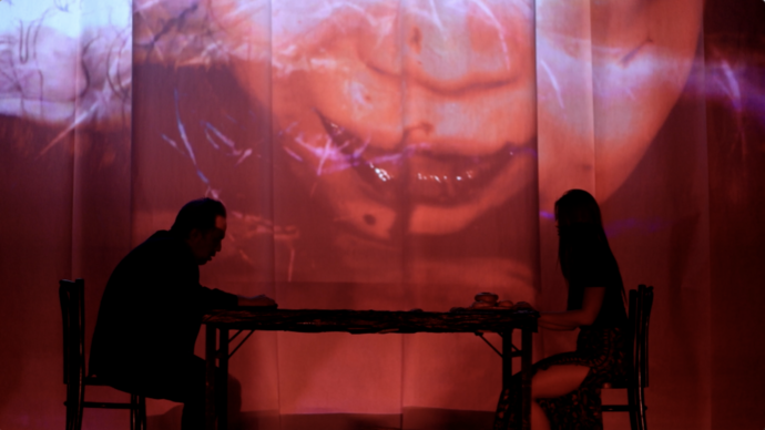 Man and woman sit at a table; distorted face projected behind creates a surreal atmosphere.
